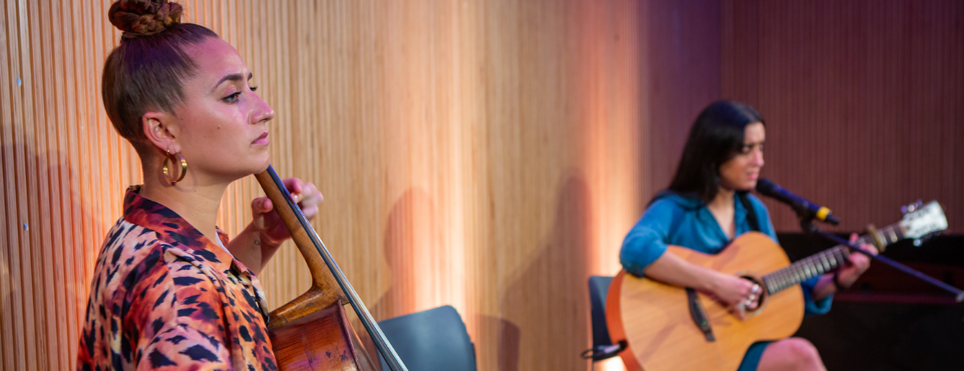 Two musicians sat on chairs on a lit stage. The musician closest to the camera is playing a cello. The musician furthest away is playing the guitar and singing into a microphone.
